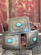A western-style belt with large engraved silver-tone square buckles, each featuring a round turquoise stone, resting on a rustic wooden surface with a blurred red decorative background.