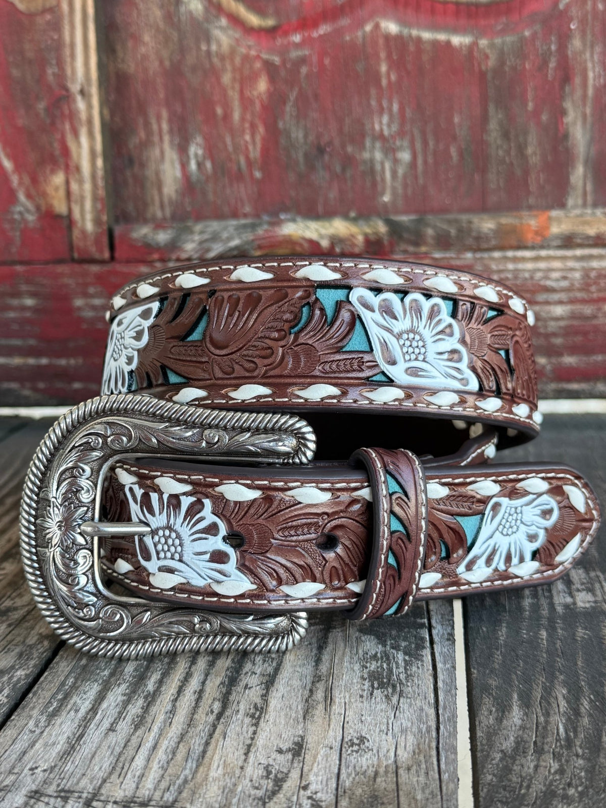 A tooled leather belt with floral cutouts and silver tooled buckle, shown against a rustic red and gray wooden background.