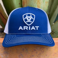 A blue and white cap featuring the white embroidered Ariat logo on the front, set against a background of green foliage and a rustic wooden surface.