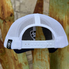 White mesh trucker cap viewed from the back, featuring a white plastic adjustment strap with 'RICHARDSON' printed on it, a black brand patch on the side, and a dark interior lining, set against a rustic wooden background.