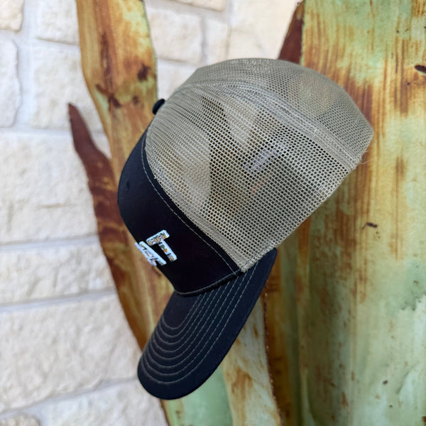A trucker-style baseball cap with a black curved brim and mesh back, featuring a small embroidered logo on the front panel, photographed against stone wall background.