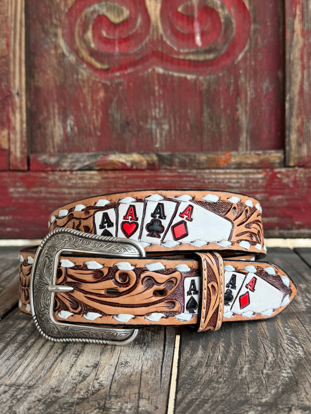 A tooled leather belt with a silver buckle, featuring a western design with white stitching and printed playing card motifs including four aces, displayed on a rustic wooden surface in front of a red wooden door.