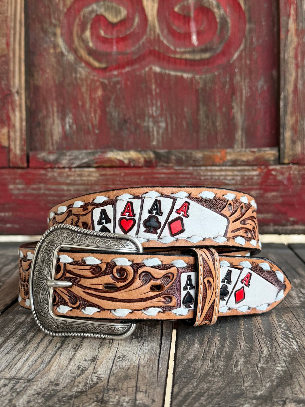 A tooled leather belt with a silver buckle, featuring a western design with white stitching and printed playing card motifs including four aces, displayed on a rustic wooden surface in front of a red wooden door.