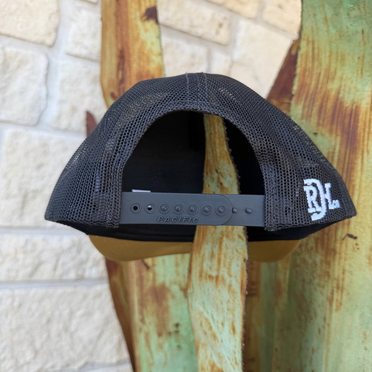 A black mesh-back trucker cap with a black adjustable strap and a white embroidered 'RDH' logo on the side, displayed on a rustic wooden stand against a light stone wall.