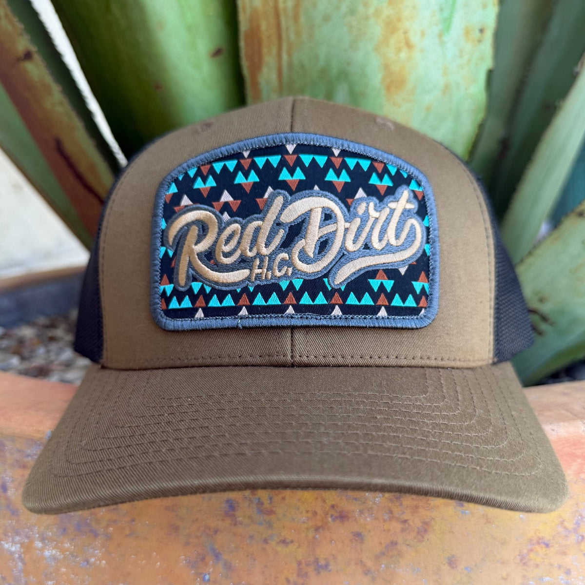 A cap with a curved brim and a front patch reading 'Red Dirt HC' in beige script over a turquoise, black, and brown geometric pattern.