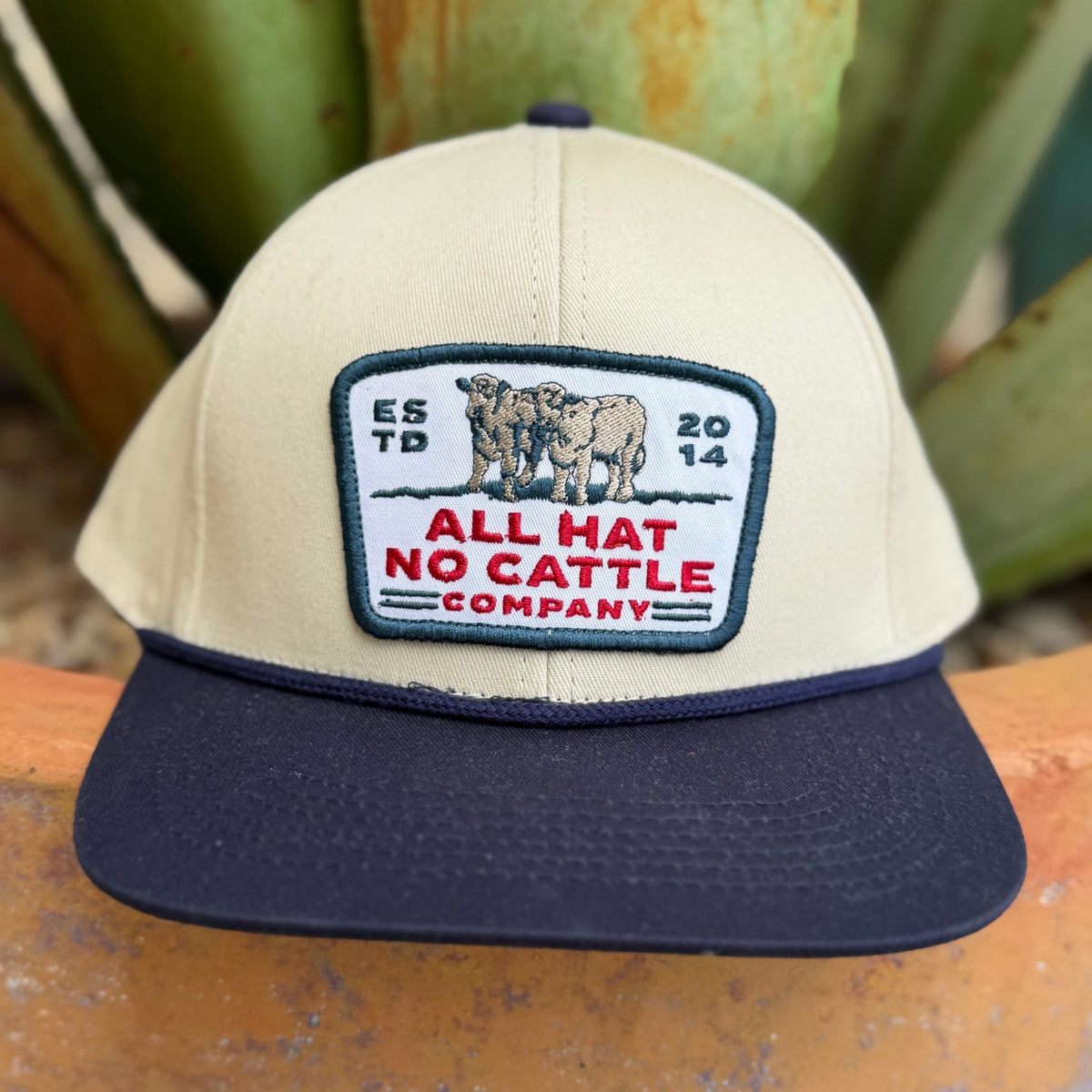 A beige and navy cap with a rectangular embroidered patch on the front that reads 'ALL HAT NO CATTLE COMPANY' with two cows and the years 'EST. 2014'.