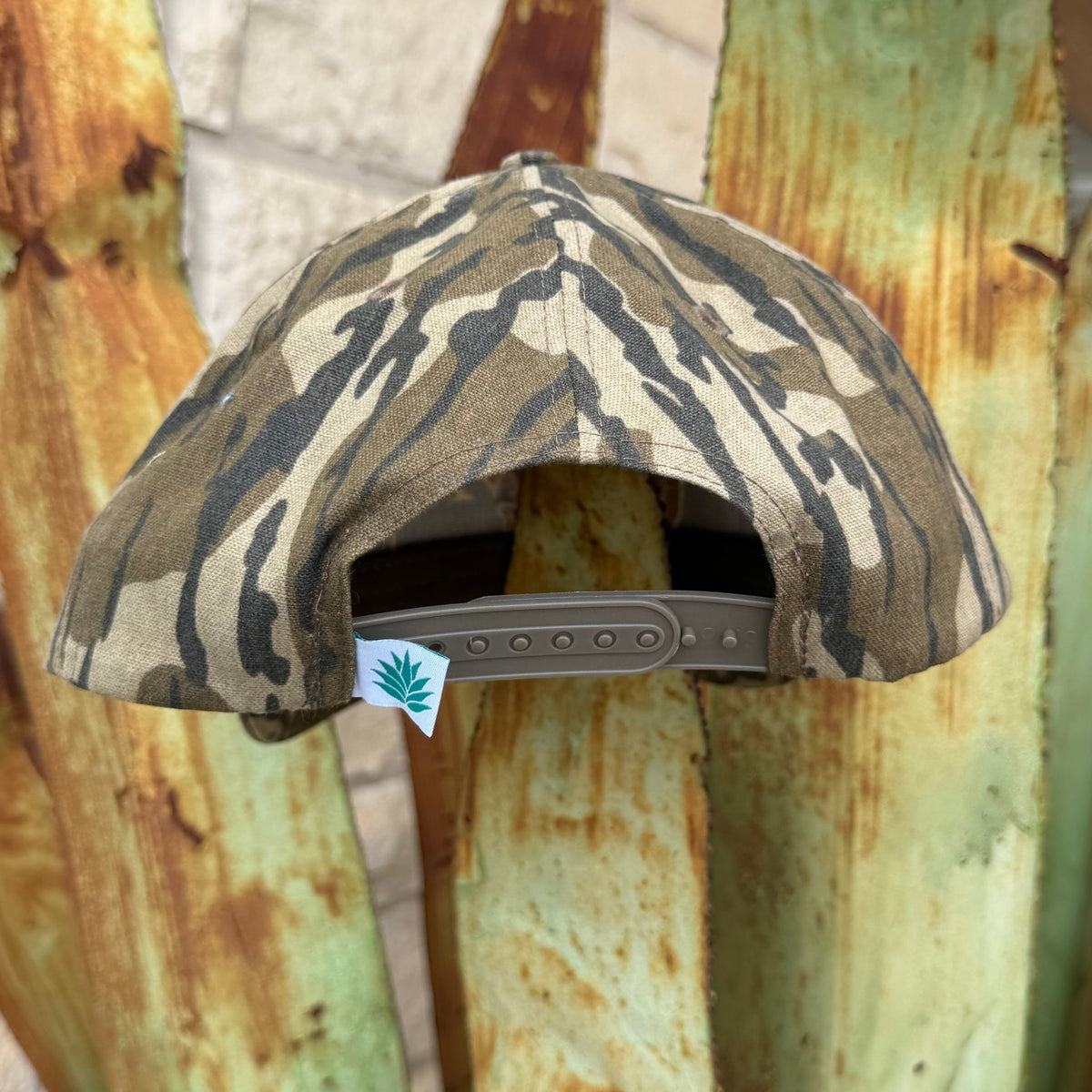 A camouflage cap with a green adjustable strap, shown from the back. The cap features a green, brown, and beige camo pattern, and a small white tag with the Sendero logo 