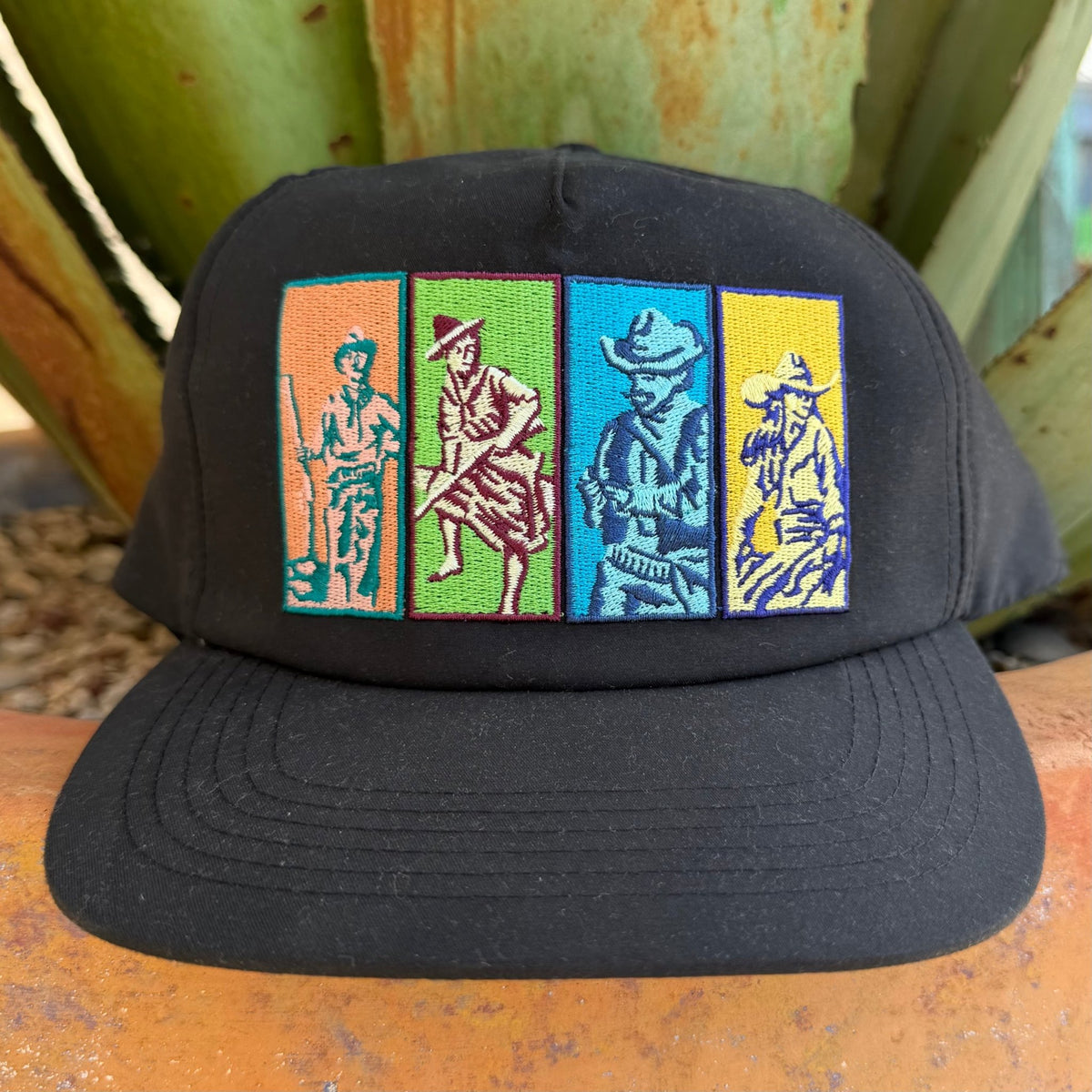 A black snapback cap featuring four colorful embroidered vertical panels with stylized cowboy and farmer figures,