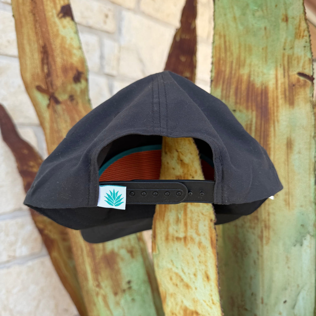 A black snapback cap shown from the back,  The back strap features a small white square patch with the Sendero Logo 