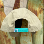 A beige cap viewed from the back, featuring a turquoise snapback closure with a small white square patch printed with the Sendero logo 