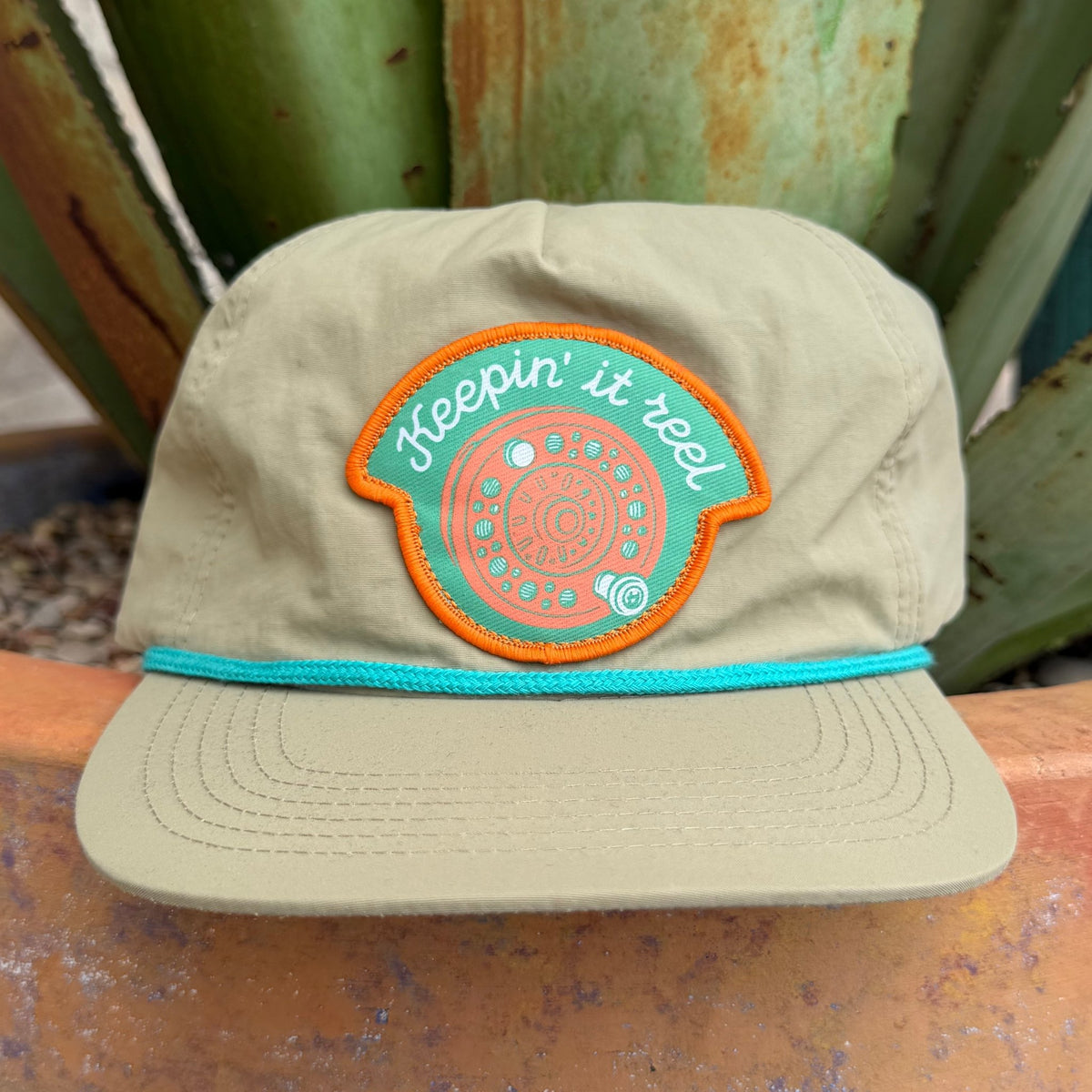 A khaki-colored cap with a turquoise and orange embroidered patch that reads 'Keepin' it reel' on the front, 