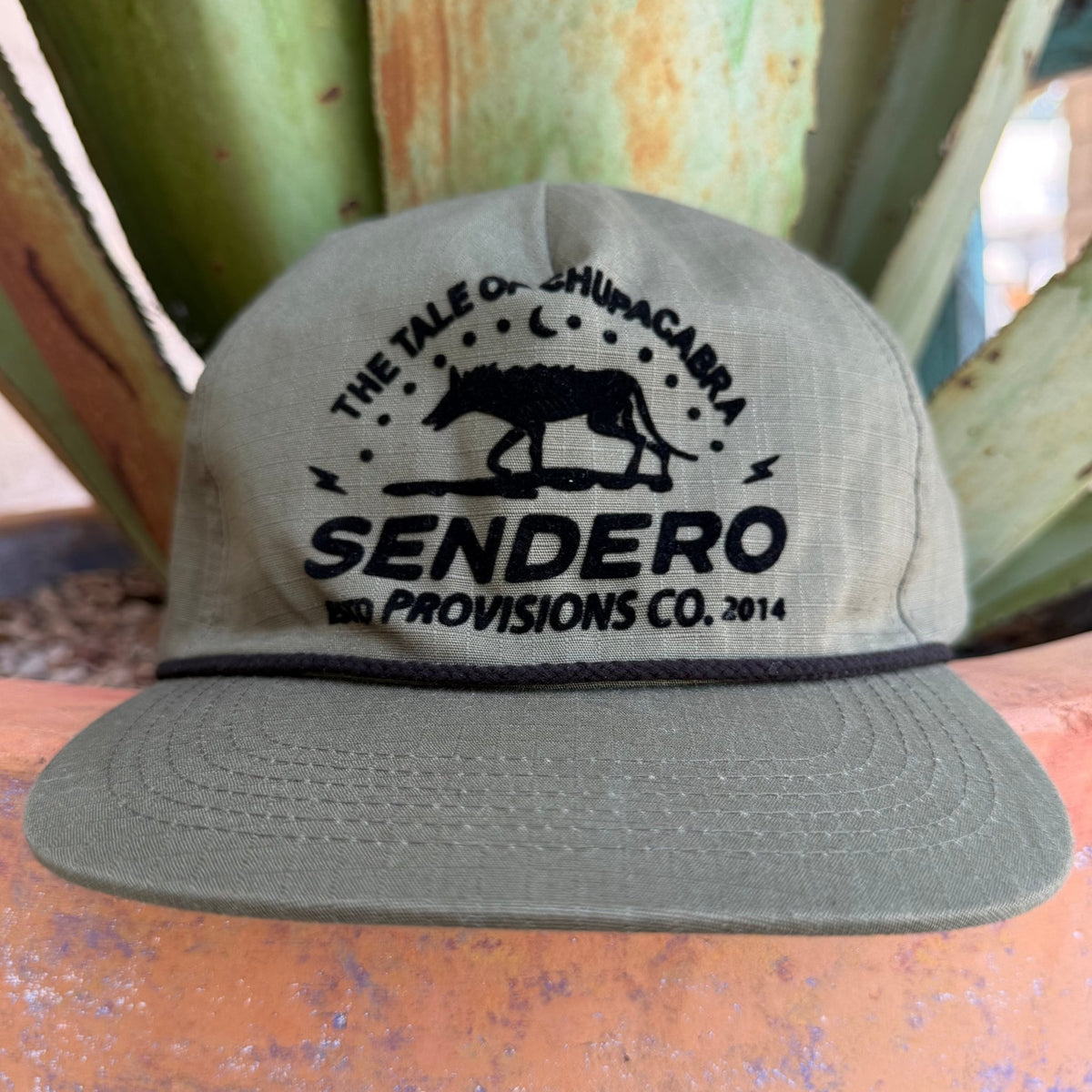 An olive green cap with a black embroidered graphic reading 'THE TALE OF CHUPACABRA SENDERO PROVISIONS CO. 2014' and a chupacabra silhouette
