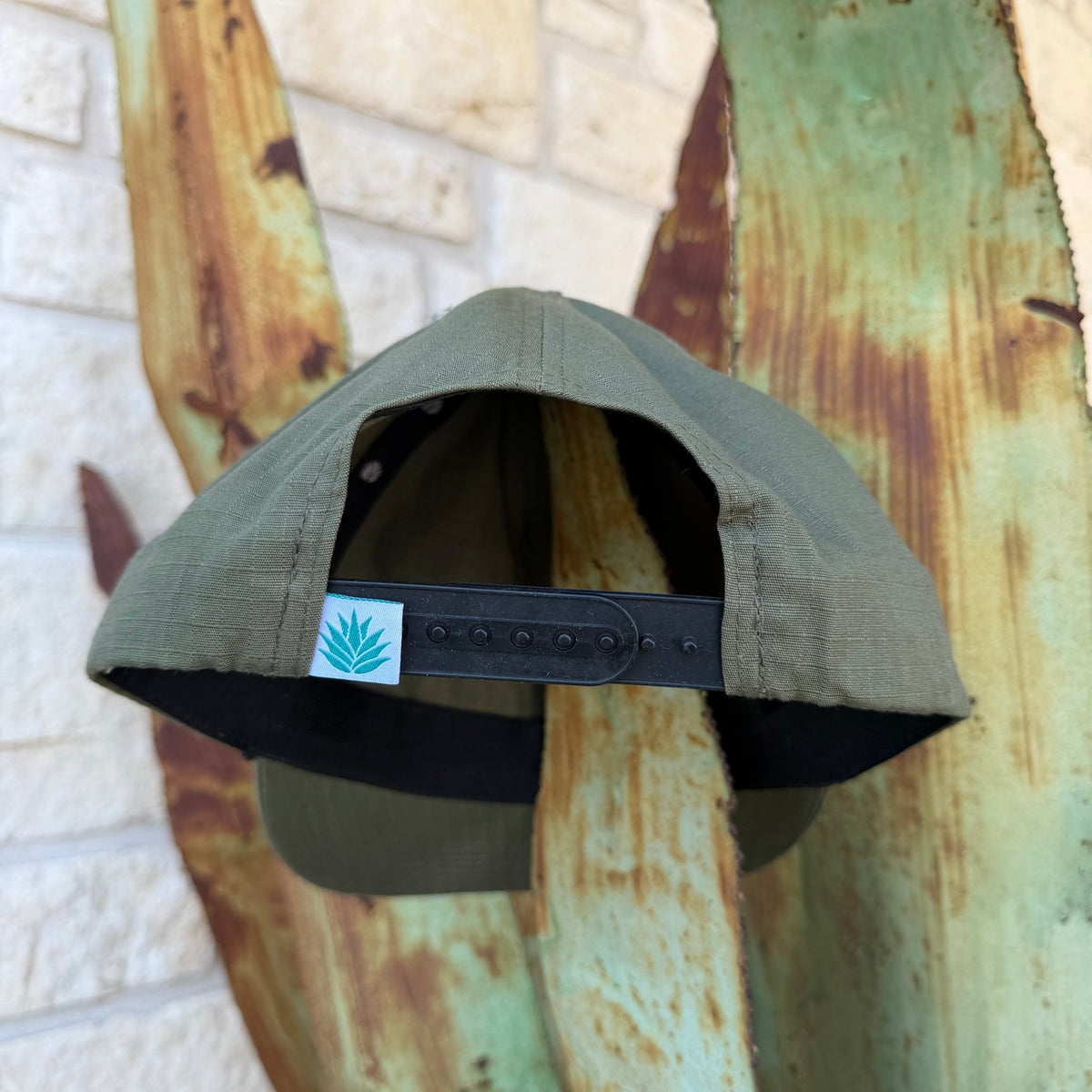 An olive green snapback cap shown from the back, featuring a black adjustable strap and a small white square patch with the Sendero logo 