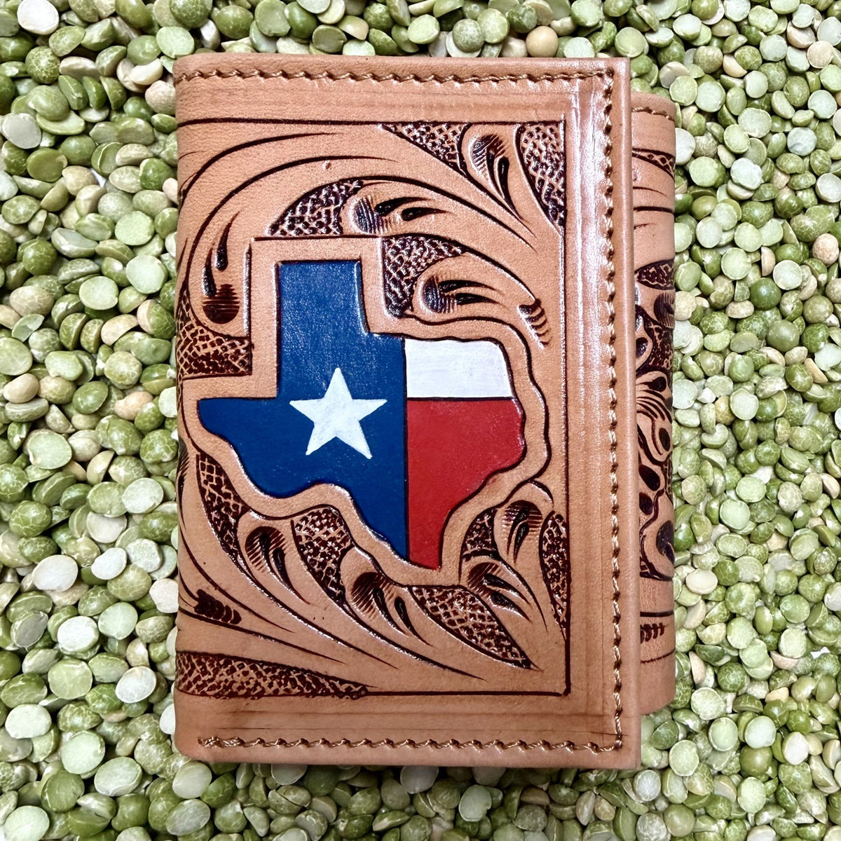 Natural tan leather trifold wallet with a Texas-shaped red, white, and blue flag accent, white star, dark floral-scroll tooling, and stitched borders — Blair's Western Wear Marble Falls TX
