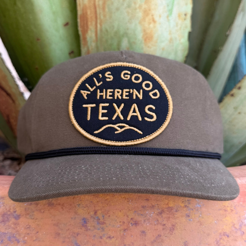 A dark olive green trucker-style cap with a circular embroidered patch reading 'ALL'S GOOD HERE'N TEXAS' in tan thread