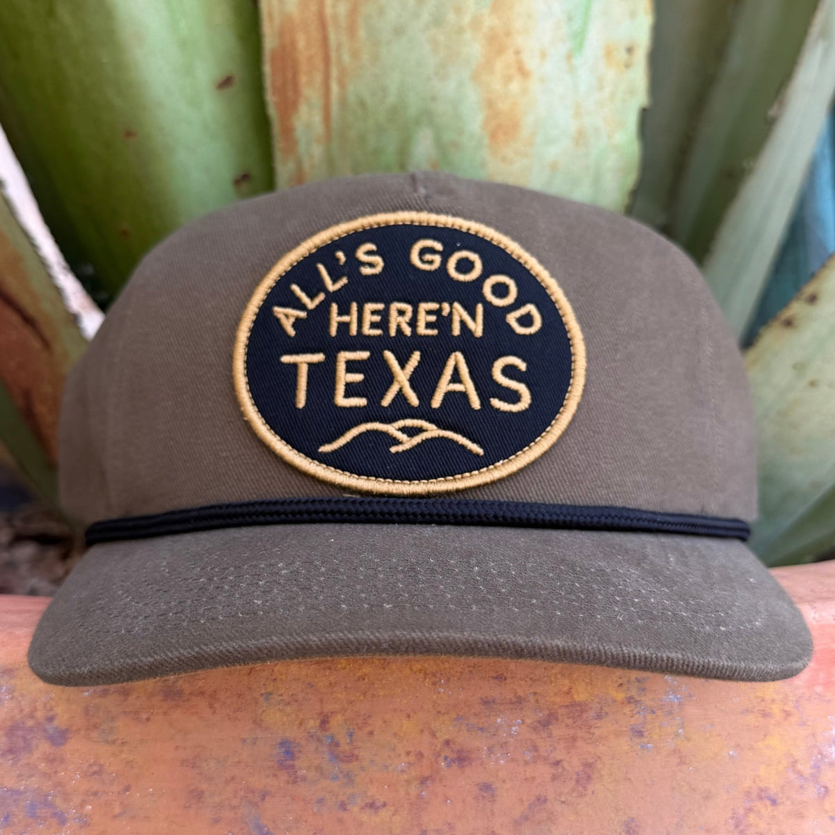 A dark olive green trucker-style cap with a circular embroidered patch reading 'ALL'S GOOD HERE'N TEXAS' in tan thread