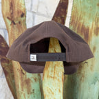 A brown structured snapback cap shown from the back. The back strap has a small white square patch with a black 'TX' logo.
