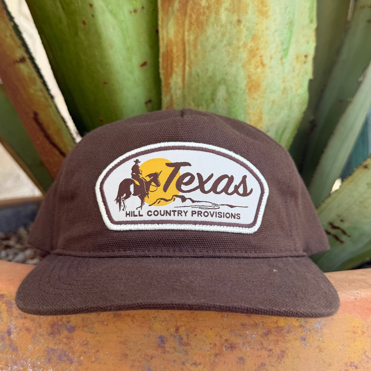 A dark brown cap with a white and yellow embroidered patch on the front that reads 'Texas Hill Country Provisions' with a cowboy and horse graphic
