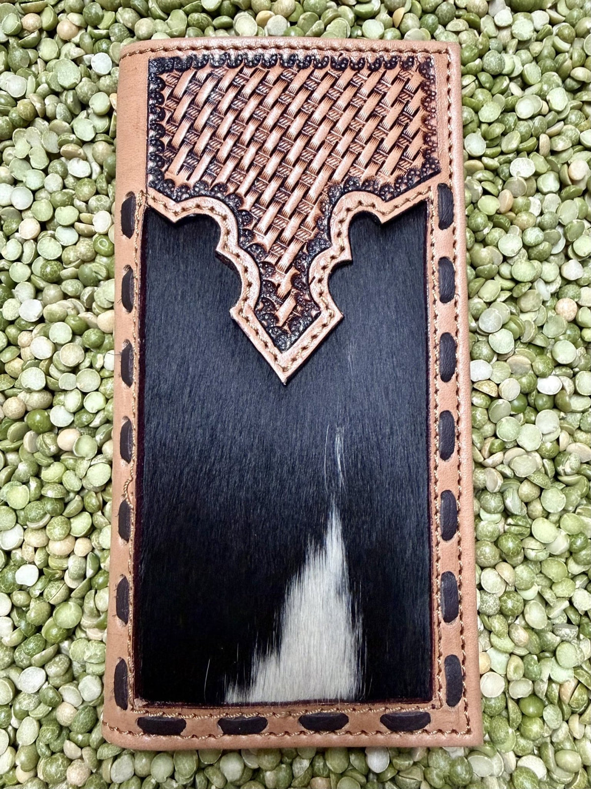 A rectangular leather wallet or card holder with a tan woven-texture top panel, decorative cutout border, and a black-and-white cowhide center, photographed from above on a bed of green lentils.