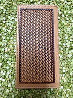 A rectangular tan leather wallet or clutch with a large woven brown and black geometric pattern panel on the front, photographed from above on a bed of green lentils.