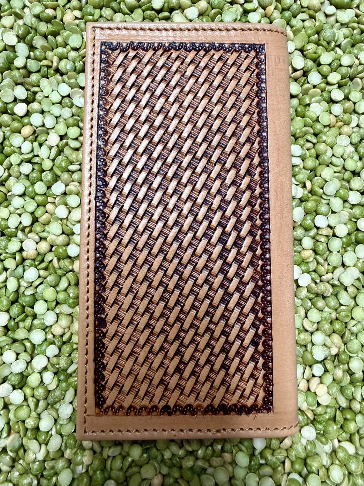 A rectangular tan leather wallet or clutch with a large woven brown and black geometric pattern panel on the front, photographed from above on a bed of green lentils.