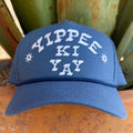 A blue baseball cap with 'YIPPEE KI YAY' embroidered in white on the front, flanked by two white starburst motifs