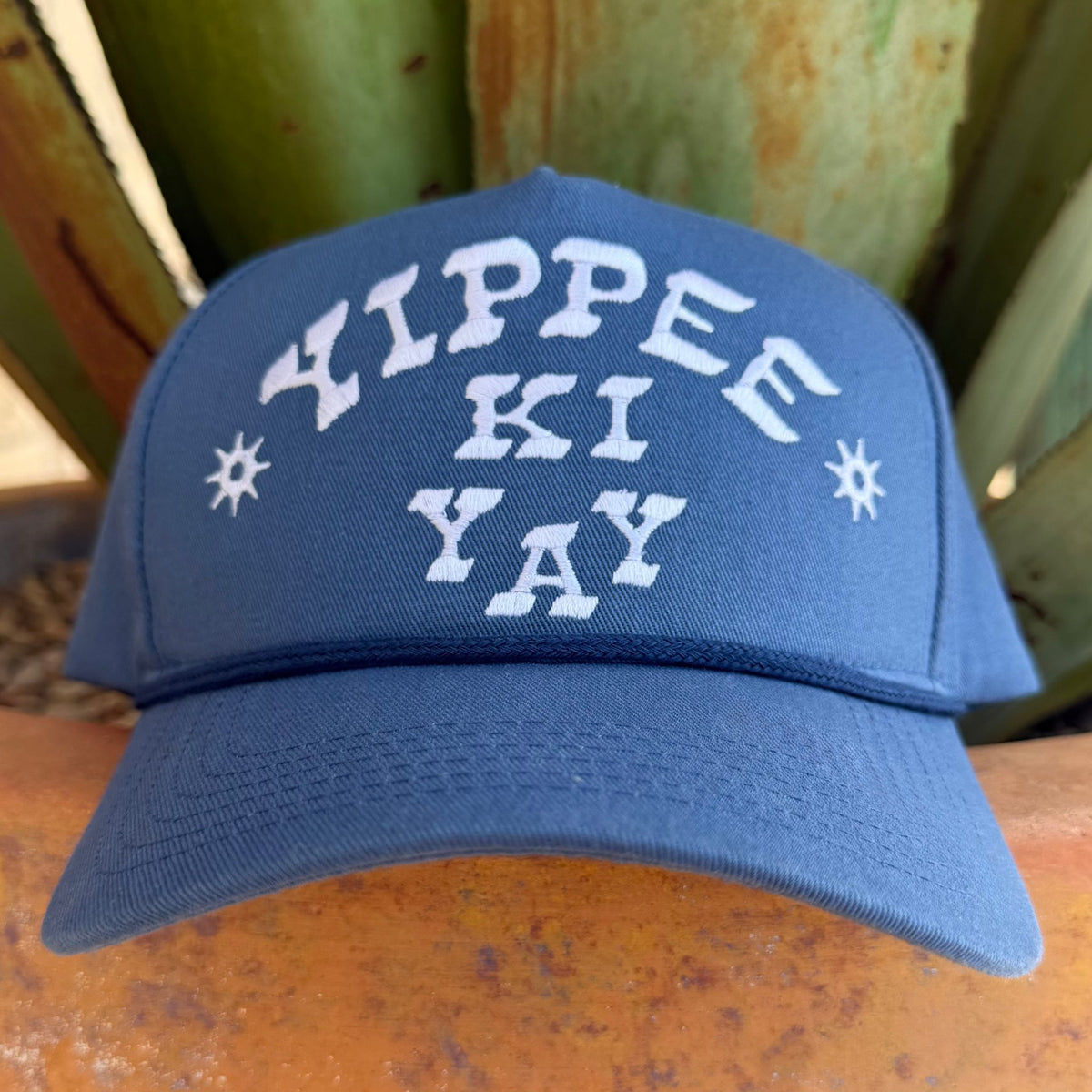 A blue baseball cap with 'YIPPEE KI YAY' embroidered in white on the front, flanked by two white starburst motifs