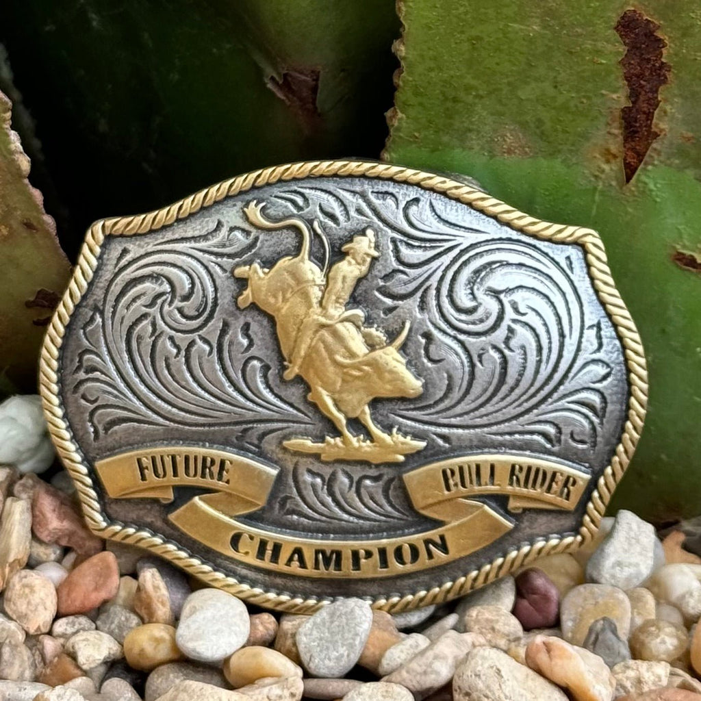 Boys’ Montana Silversmiths buckle in antique silver and gold with a central bull rider figure, engraved floral detailing, rope-edge border, sturdy zinc alloy construction, and secure 1.5″ swivel hook – A1025P. Available at Blair's Western Wear, Marble Falls, TX.