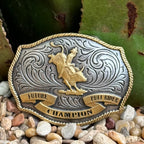 Boys’ Montana Silversmiths buckle in antique silver and gold with a central bull rider figure, engraved floral detailing, rope-edge border, sturdy zinc alloy construction, and secure 1.5″ swivel hook – A1025P. Available at Blair's Western Wear, Marble Falls, TX.
