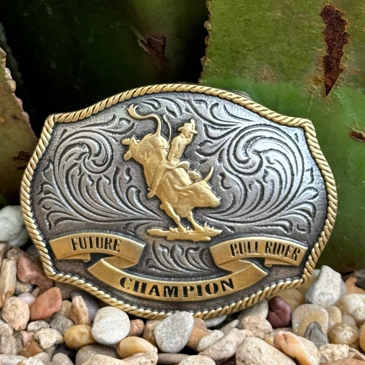 Boys’ Montana Silversmiths buckle in antique silver and gold with a central bull rider figure, engraved floral detailing, rope-edge border, sturdy zinc alloy construction, and secure 1.5″ swivel hook – A1025P. Available at Blair's Western Wear, Marble Falls, TX.