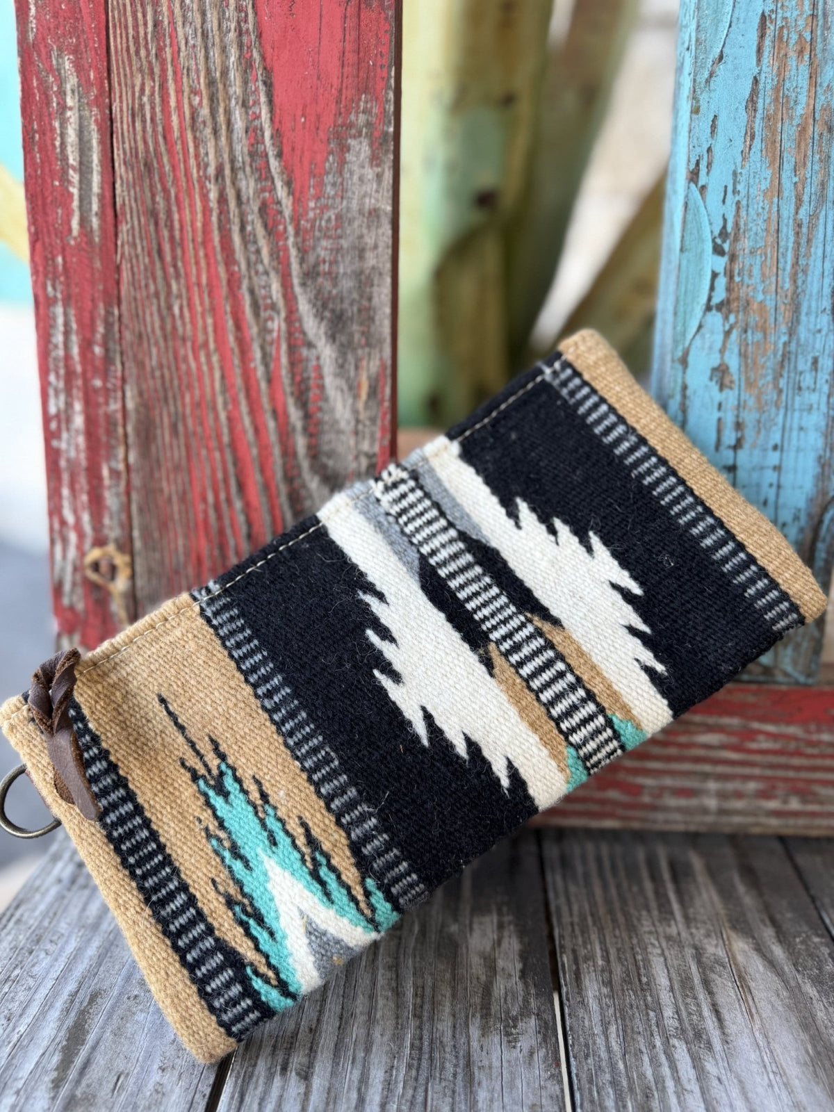 ADBG344BC — Front view of women's black, cream, and turquoise Navajo-inspired blanket wristlet showing bold geometric saddle blanket pattern with cream/white lightning bolt motifs, turquoise accents, houndstooth checker border stripes, dark brown leather zipper pull at top left, and brown leather wrist strap with silver D-ring and snap on the right — Blair's Western Wear Marble Falls TX