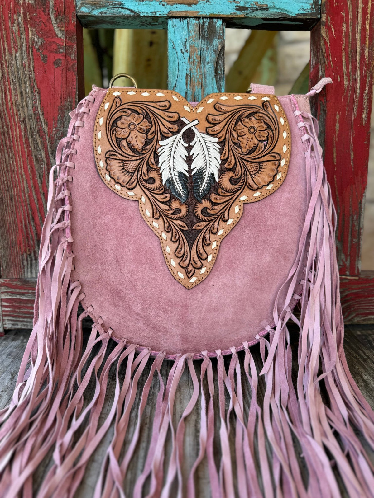 ADBGA431A — This dreamy blush pink suede crossbody is pure boho western romance — long blush fringe on both sides and bottom, a sculpted heart-shaped tan hand-tooled floral leather flap with a single large white and dark gray feather centerpiece, gold diamond whipstitch trim, and a brass D-ring at the top — Blair's Western Wear Marble Falls TX