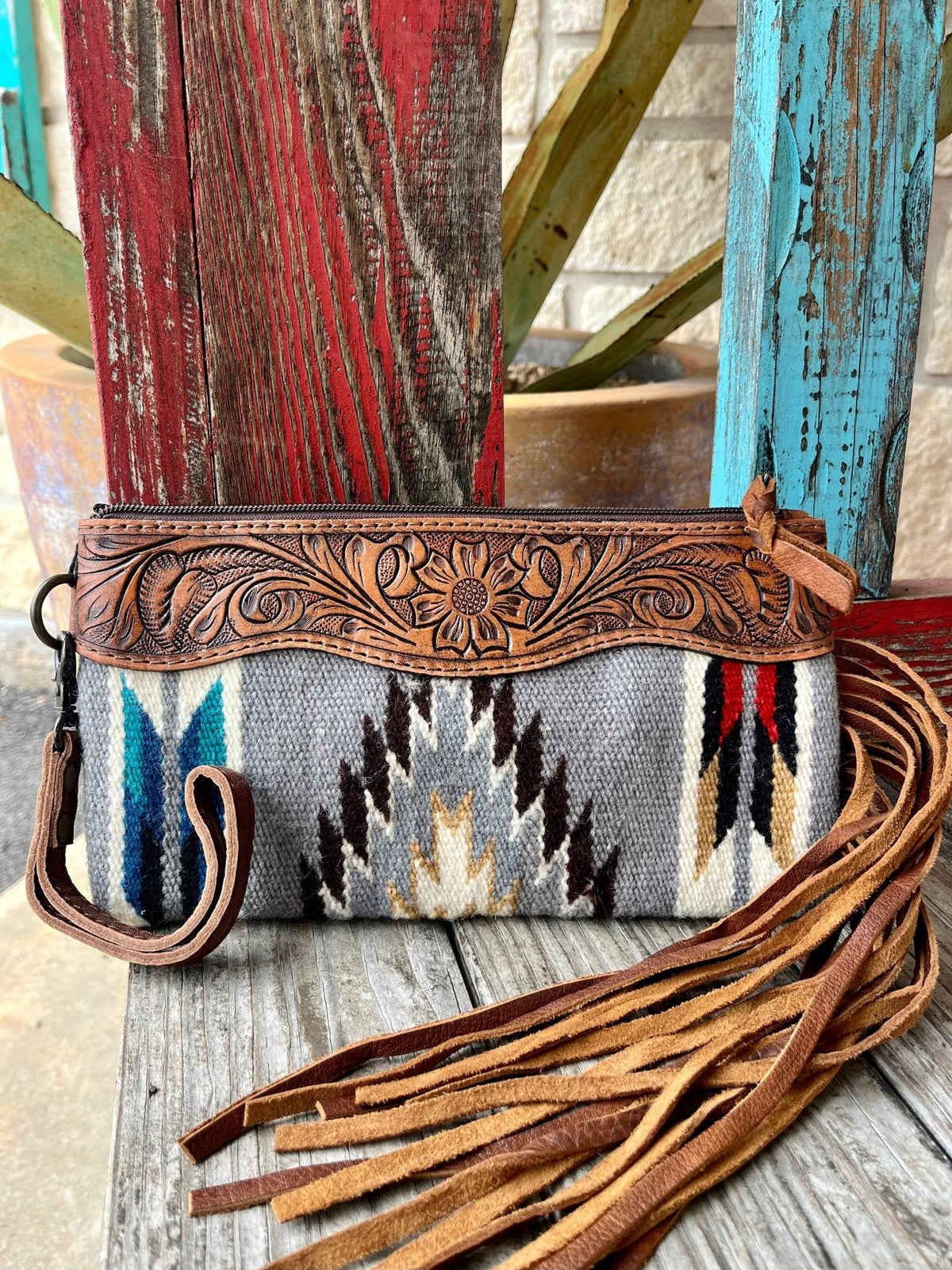 ADBG344D4 — Women's grey Aztec-inspired blanket wool wristlet with bold feather and arrow geometric pattern in turquoise/teal, navy, white, black, chocolate brown, red, and tan/gold, wide medium brown hand-tooled leather top yoke featuring central sunflower medallion with sweeping acanthus scroll and leaf tooling and scalloped bottom edge, long tan/caramel suede leather fringe on the right side, gunmetal zipper with leather pull tab, and gunmetal swivel clip wristlet strap on the left — Blair's Western Wear