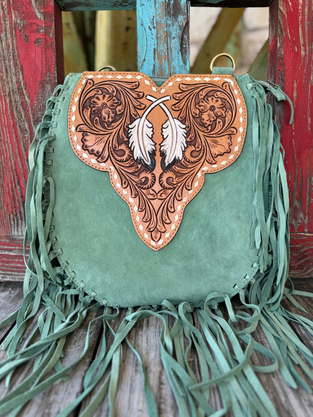 ADBGA431D — Angled view of women's sage green suede fringe crossbody showing sculpted hand-tooled floral feather flap with two white painted feathers and cream whipstitch trim, with the tan hand-tooled feather-scroll crossbody strap and antique brass swivel hardware displayed in front — Blair's Western Wear Marble Falls TX