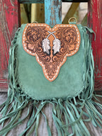 ADBGA431D — Angled view of women's sage green suede fringe crossbody showing sculpted hand-tooled floral feather flap with two white painted feathers and cream whipstitch trim, with the tan hand-tooled feather-scroll crossbody strap and antique brass swivel hardware displayed in front — Blair's Western Wear Marble Falls TX