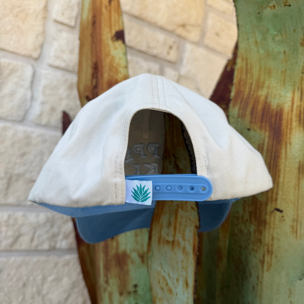 A light beige snapback cap with a blue visor and blue adjustable strap, featuring a small green aloe vera patch on the side.