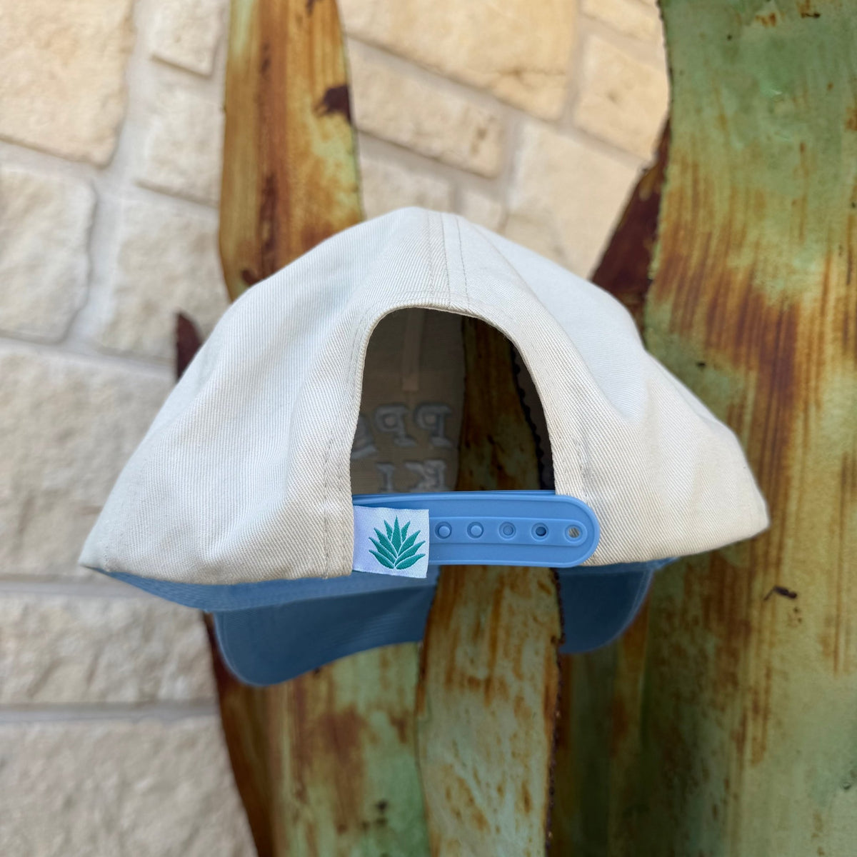 A light beige snapback cap with a blue visor and blue adjustable strap, featuring a small green aloe vera patch on the side.