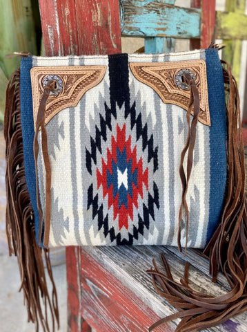 ADBGA289H — Women's large Navajo-inspired wool tote with cream/white, gray, and navy hand-woven wool body featuring bold Navajo-inspired geometric star and diamond medallion in navy, red, turquoise, and white, cobalt blue woven side panels, pale natural tan hand-tooled leather triangular corner yokes with floral scroll tooling and silver concho snap hardware, and long dark brown leather fringe on both sides — Blair's Western Wear Marble Falls TX